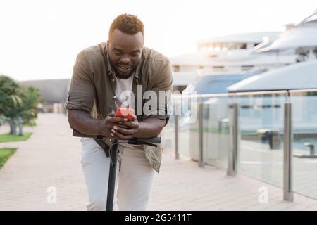 Young african man having fun driving electric scooter while using smartphone during summer vacation - Eco travel concept Stock Photo