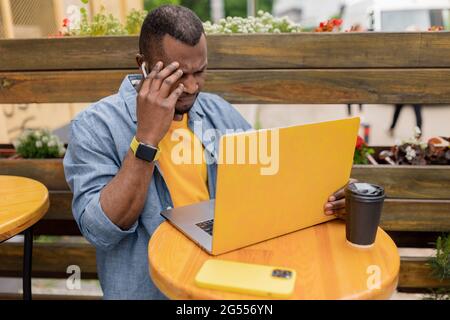 Frustrated overworked afro american man hold online conference or checking email on laptop pc in outdoor cafe take away, receiving bad news, touching Stock Photo
