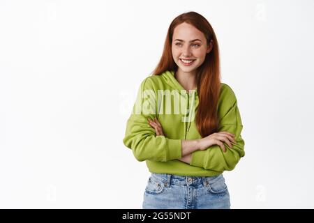 Cheerful ginger girl laughing, smiling and looking carefree aside, standing relaxed pose with crossed arms, wearing green hoodie and jeans, white Stock Photo
