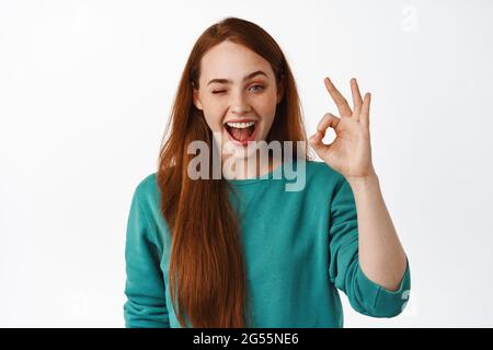 Cheerful ginger girl assure you, say no zero problems, winking and smiling, show okay OK gesture, standing pleased, leave positive feedback, white Stock Photo