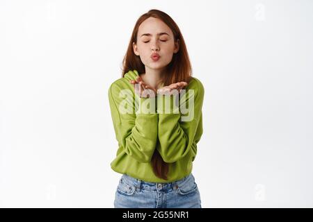 Portrait of tender redhead woman kissing, sending air kiss in open hands, close eyes and standing romantic against white background, isolated shot Stock Photo