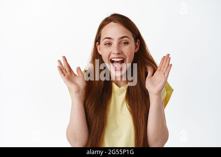 Close up of happy laughing redhead woman, clap hands and smiling amazed, checking out awesome promo offer, great news, standing against white Stock Photo