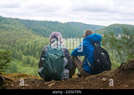 two teenagers with backpacks are sitting on the edge of a cliff, looking into the distance to the wooded hills Stock Photo