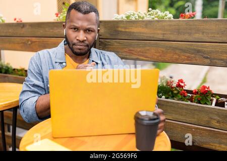 Afro American focused pensive male looking at camera, working on laptop in cafe outdoors, enjoying distant work after quarantine.Stylish millennial Stock Photo