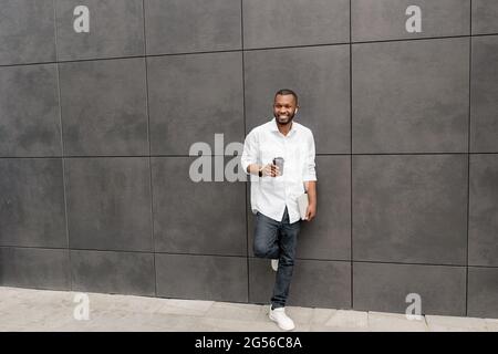 Stylish smiling Afro American millennial freelancer or student in casual clothes having break after hard day, drinking take away coffee Stock Photo