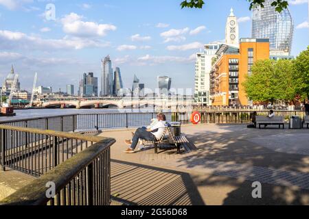 London, England - 22 May, 2019: People sitting on a bench by the river bank of River Thames near Thames Beach with office towers in the background. Stock Photo