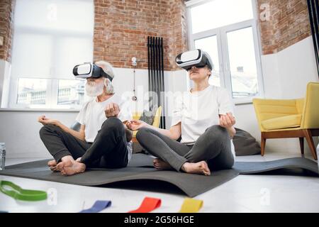 Relaxed aged man and woman wearing VR glasses while sitting in lotus position at bright living room. Active senior couple meditating at home using innovative headset. Stock Photo