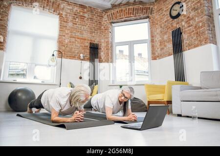 Strong retired couple keeping balance on yoga mat while doing plank exercise. Active man and women in sport outfit watching online tutorial on laptop. Stock Photo