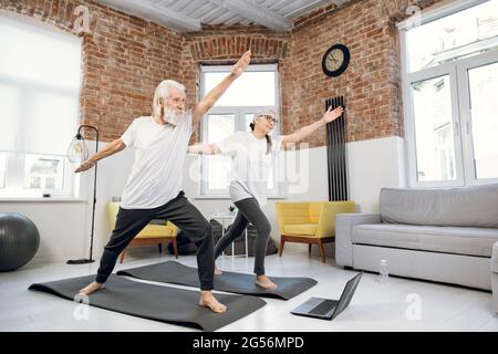 Two elderly people in sport clothes standing in yoga asana pose while watching online tutorial on modern laptop. Family training at home. Active retirement. Stock Photo