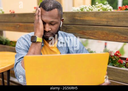 Pensive concentrated Afro American teacher having headache, sitting at table in cafe outdoors, working remotely, holding online lessons with students Stock Photo