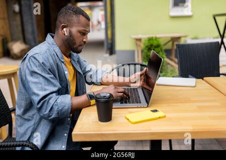 Side view of focused pensive Afro American freelancer wear wireless headphones, holding online meeting, watching educational lessons on laptop Stock Photo