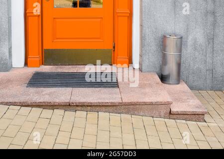 granite threshold with foot mat near orange wooden front door with iron trash can near building facade with gray cladding and yellow stone tile paveme Stock Photo