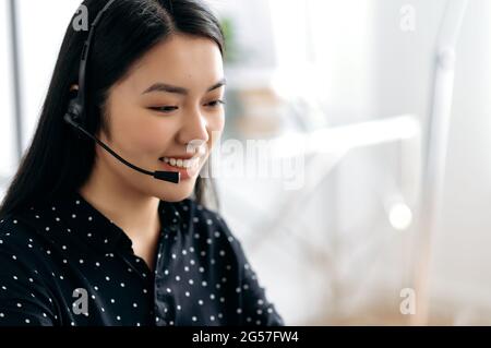 Side view of a pleasant, satisfied young asian woman with headset, freelancer, call center worker or consultant, hotline operator, smiles friendly, conducts online consultation, talks with client Stock Photo