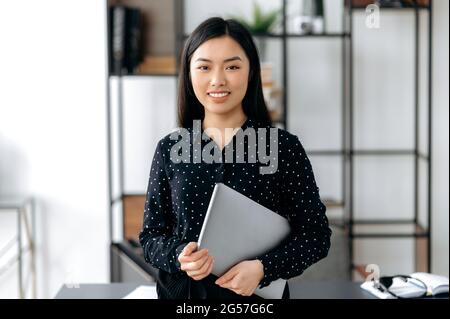 Portrait of a confident, pretty, young japanese or chinese woman. Female asian girl, office manager, stand near work desk in office, holds laptop in hands, looks at camera, smiles friendly Stock Photo