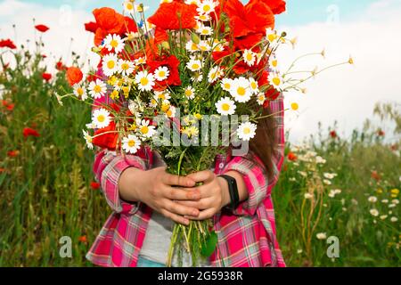 The girl stands against the background of wild flowers and holds a bouquet of poppies and daisies in her hands. A girl in the field was collecting a bouquet of wild flowers. Stock Photo