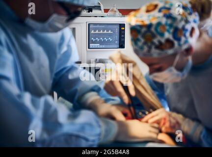 Focus on heart rate monitor and medical team performing tummy tuck surgery in operating room. Plastic surgeon and assistant removing excess fat from patient abdomen. Concept of abdominoplasty. Stock Photo