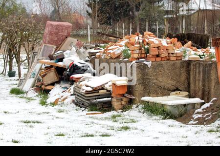 Bunch of various junk from construction material made of bricks and bathtub to broken windows and old sofa next to dilapidated family house concrete Stock Photo