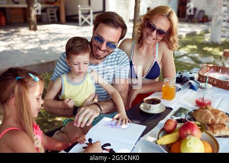sister  showing to a brother how to draw at outdoor table, having breakfast with parents Stock Photo