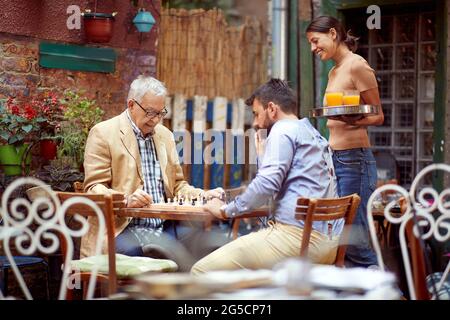 two men, young and senior, playing chess in outdoor cafe, thinking, with waitress beside watching and smiling. selective focus image with copy space Stock Photo