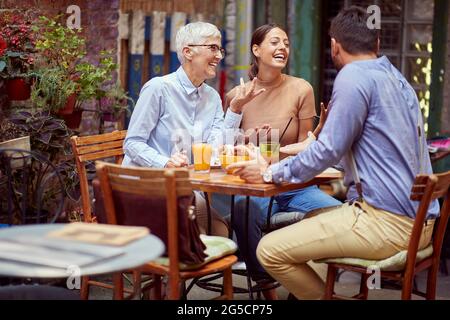 elderly woman  having conversation with young adult female and her boyfriend, smiling, having breakfast together in outdoor cafe. family communication Stock Photo