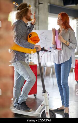 A young male employee on a scooter is having fun in a friendly atmosphere in the office while chatting with his female colleague Stock Photo
