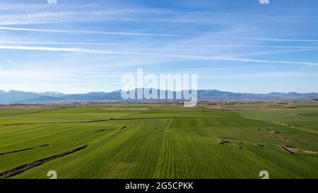 Spectacular aerial view of a green meadow with a city in the background at the foot of a mountain range silhouetted against a blue sky dusted with ten Stock Photo