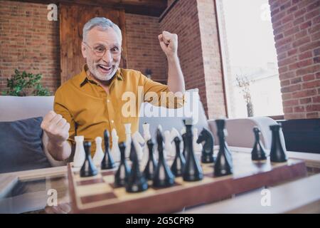 Photo of old man playing chess celebrating succsessful step raising fists player home indoors Stock Photo