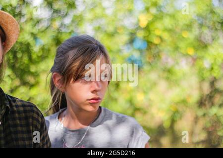 Defocused close-up preteen serious blond caucasian girl. Summer blurred green nature bokeh background. Portrait of handsome confused girl. Out of Stock Photo