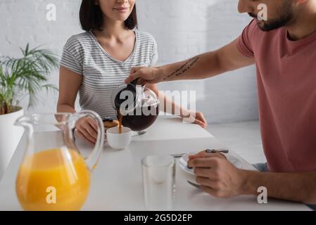 partial view of young man pouring coffee from pot to cup near girlfriend sitting at table with breakfast in kitchen Stock Photo