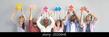 Group of happy young people holding pieces of jigsaw puzzle standing against light background Stock Photo