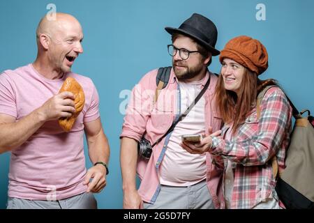 Tourists, a smiling woman with a smartphone and an incredulous man ask the route from a cheerful local who holds a pie in hands.  Stock Photo