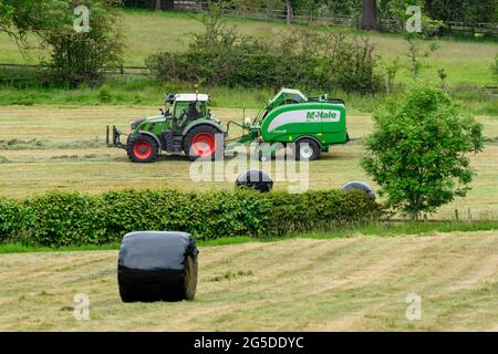 Hay or silage making (farmer in farm tractor at work in rural field pulling baler, collecting dry grass & round bales wrapped - Yorkshire England, UK. Stock Photo