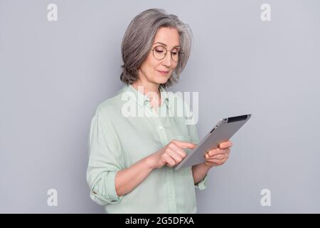 Photo of positive lovely focused concentrated mature woman in glasses using tablet isolated on grey color background Stock Photo