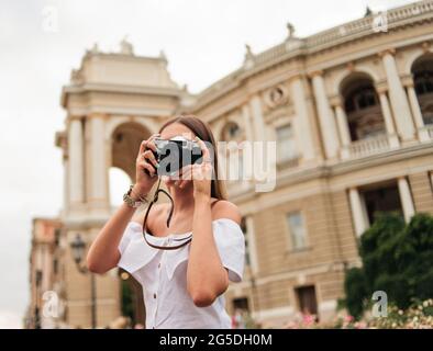 Young happy woman holds in her hands retro camera and and takes a picture against the background of the architecture of tourist city. Traveling to new Stock Photo