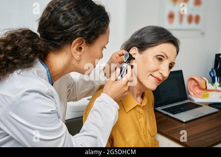 Hearing exam for elderly citizen people. Otolaryngologist doctor checking mature woman's ear using otoscope or auriscope at medical clinic Stock Photo