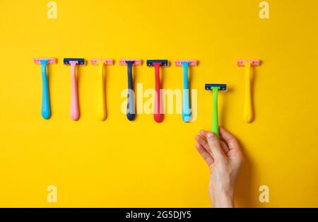 Female hand choosing green razor among a variety of colored epilator razors on yellow background. Top view. Minimalism beauty concept. Top view Stock Photo