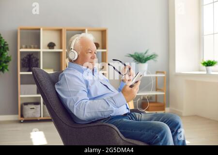 Senior man sitting in armchair at home wearing headphones and using mobile phone Stock Photo