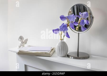 A branch of blue iris in a white fluted vase next to a round mirror, a figurine of an angel and a pile of white papers on a table against a white wall Stock Photo