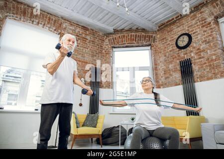 Grey haired man using dumbbells for training arms while his charming wife workout with resistance band on fitball. Two retired people doing morning exercise at home. Stock Photo