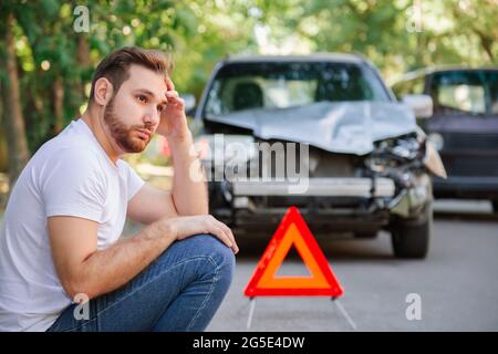 Portrait of upset man sitting on road near wrecked car after car accident with red triangle. Caucasian scary driver man sitting near his wrecked car Stock Photo