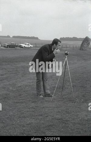 1960s, historical, a man in a jacket and tie using a camera on a tripod to take a picture of the ancient stones at Stonehenge, Wiltshire, England, UK. At this time one could walk freely around the prehistoric stones but in the following decade, in 1977, due to the increasing large numbers of visitors, damage to the grass and general erosion at the sacred, ancient burial site, the stones were roped off preventing access. On land owned at one time by King Henry VIII, the Amesbury Abbey Estate, Cecil Chubb brought the plot and the stones at an auction in 1915 and in 1918 gave it to the nation. Stock Photo
