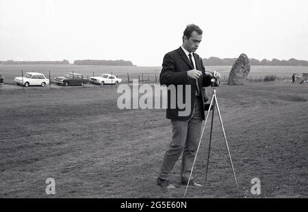 1960s, historical, a man in a jacket and tie using a camera on a tripod to take a picture of the ancient stones at Stonehenge, Wiltshire, England, UK. At this time one could walk freely around the prehistoric stones but in the following decade, in 1977, due to the increasing large numbers of visitors, damage to the grass and general erosion at the sacred, ancient burial site, the stones were roped off preventing access. On land owned at one time by King Henry VIII, the Amesbury Abbey Estate, Cecil Chubb brought the plot and the stones at an auction in 1915 and in 1918 gave it to the nation. Stock Photo