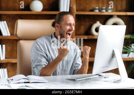 Happy excited lucky caucasian young man, stylishly dressed, office worker, freelancer, ceo sits at work desk, gesturing with his fists rejoicing in victory, success, good deal, big profit Stock Photo