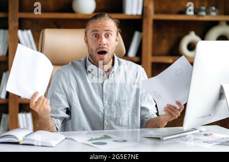 Shocked amazed caucasian guy, dressed in shirt, manager or office employee, sitting at the table, holding documents, charts in his hands, looking straight into the camera in surprise Stock Photo