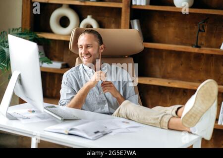 Attractive positive young adult american man, manager or ceo, sitting in office with feet on table, using computer, communicating via conference call with colleagues or friends, reading news, smiling Stock Photo