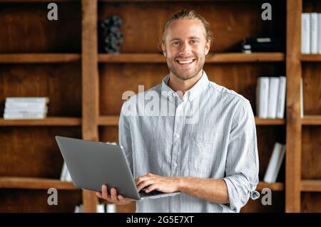 Portrait of a successful confident young man. Handsome caucasian, american guy stands in the office in a stylish formal shirt, holds an open laptop, looks at the camera and smiles friendly Stock Photo