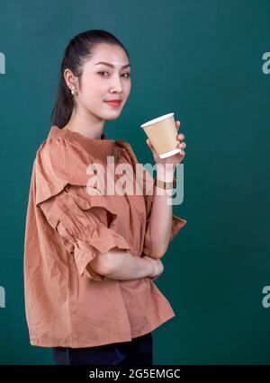 Young asian woman in casual dress with a smile holding coffee drink in paper cup. Portrait on green background with studio light.