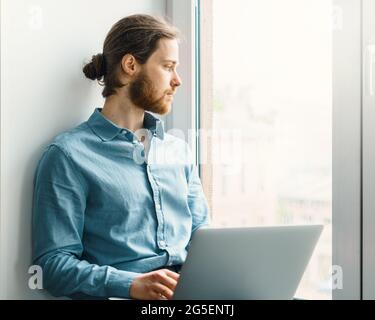 Young dreamy bearded man sitting with laptop on windowsill Stock Photo