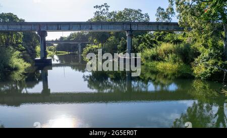 A country creek with a railway bridge and a road overpass and water reflections and forested banks in early morning sunlight Stock Photo
