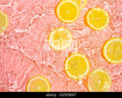 Lemon slices in clean transparent water over pink background with copy space. Water splashing on water surface in sunlight. Top view or flatlay. Summer, vacation, healthy eating concept Stock Photo